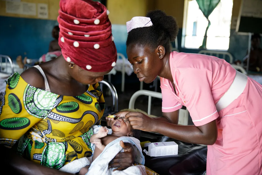 A woman holds a baby while a nurse delicately places eyedrops in the baby's eyes.