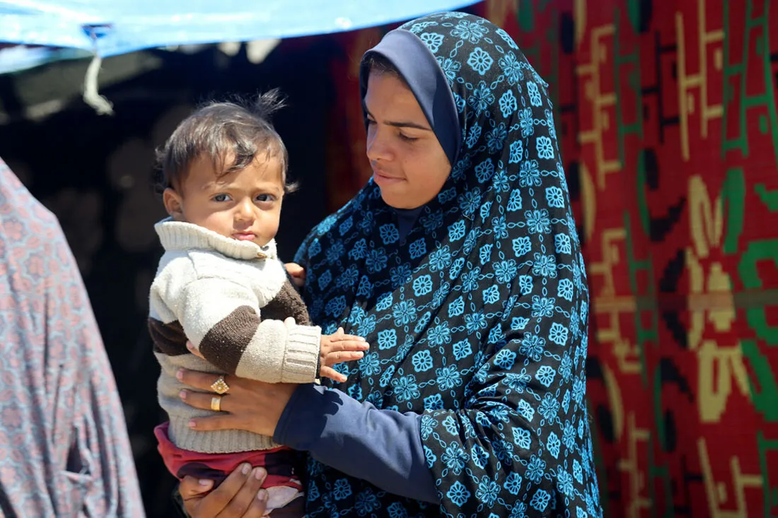 A woman holds a small child in front of a makeshift tent.