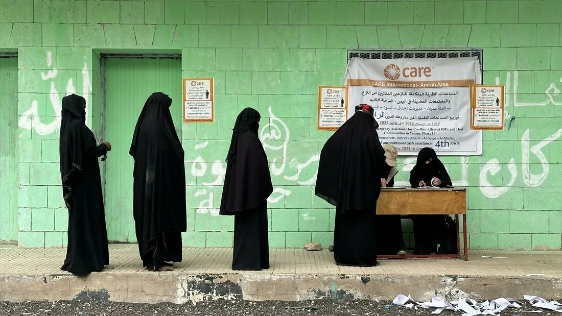 A group of women stand in single file to speak with someone at a desk.