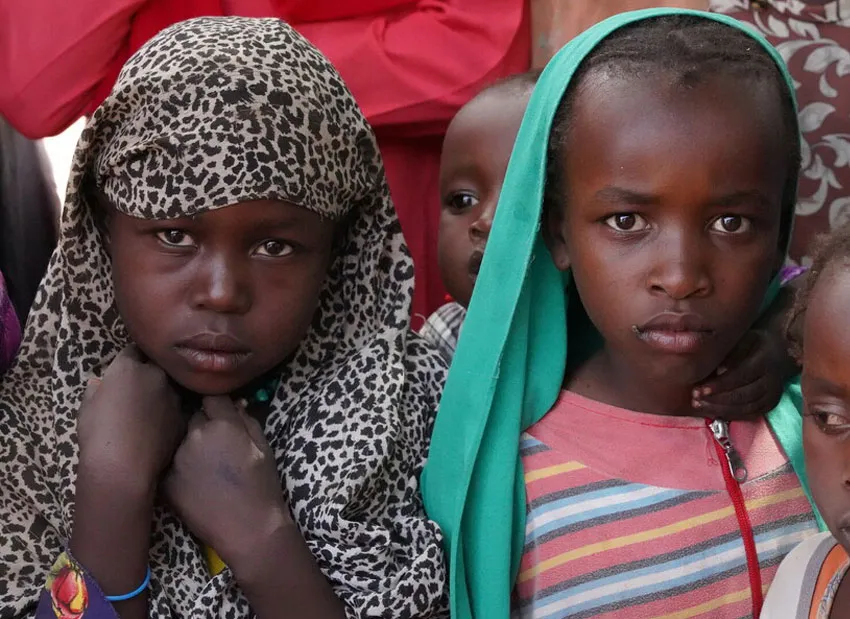 Three Sudanese children stand next to each other, one of them clutching fabric around their head.