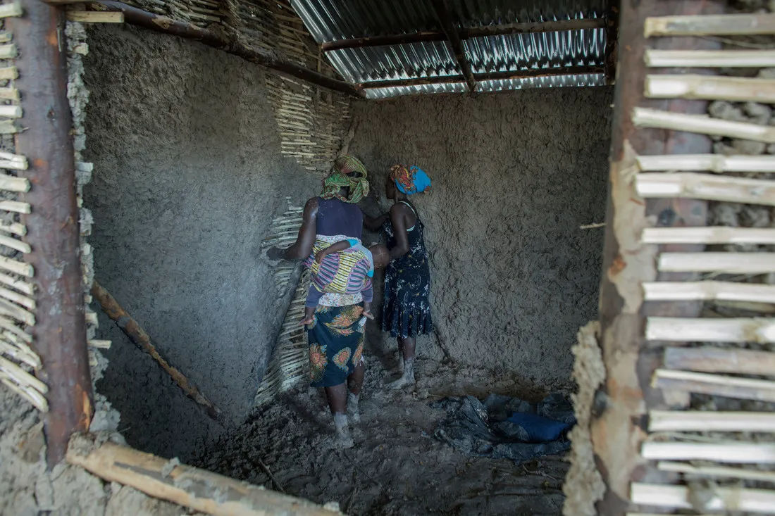 Two Zimbabwean women work together to construct or repair a mud and bamboo house. A young child is carried on the back of one of the women.