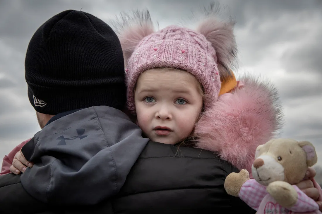 A young girl wearing a light pink winter coat and matching hat looks over the shoulder of the man carrying her.