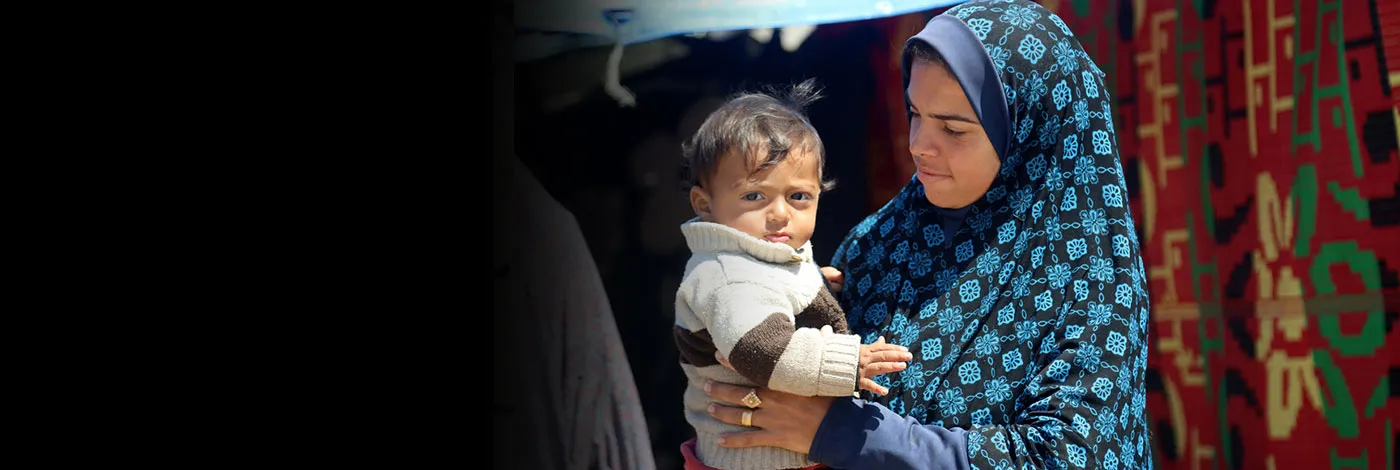 A woman holds a small child in front of a makeshift tent.