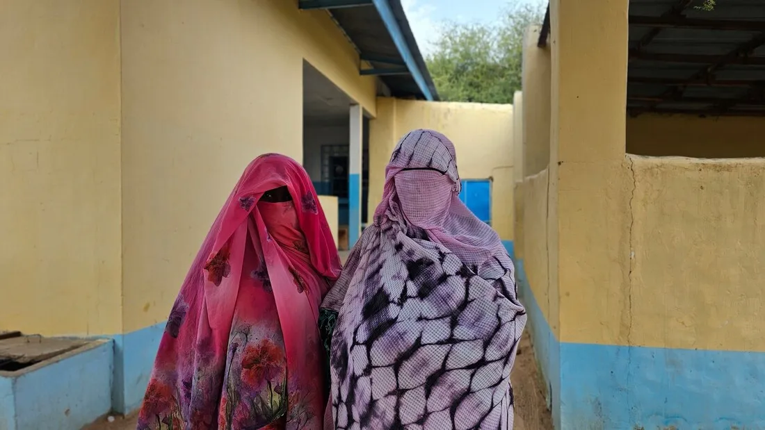 Two veiled women pose together, looking at the camera in front of a house.