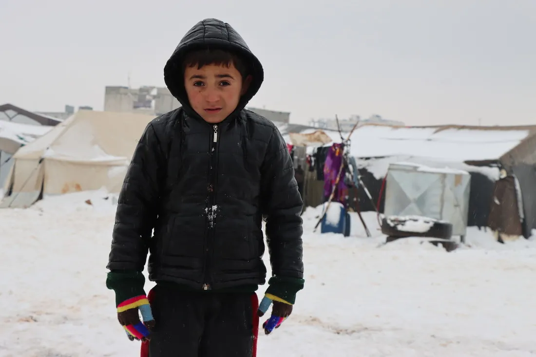 A Syrian boy stands outside in the snow in a light jacket and gloves.