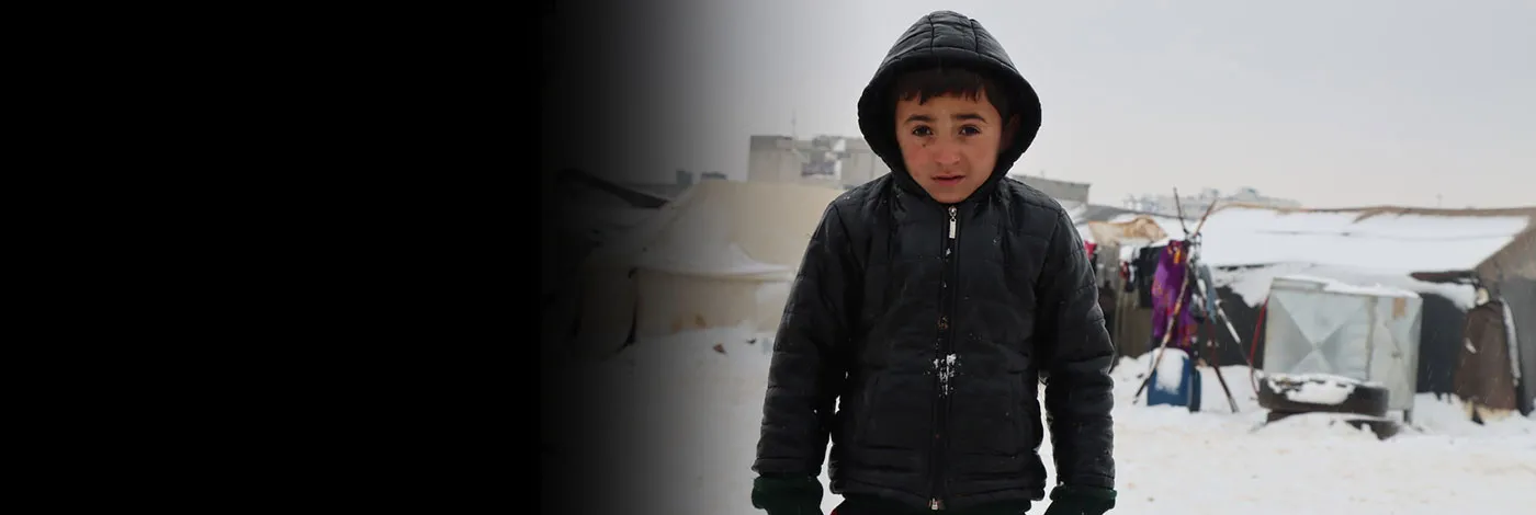 A Syrian boy stands outside in the snow in a light jacket and gloves.